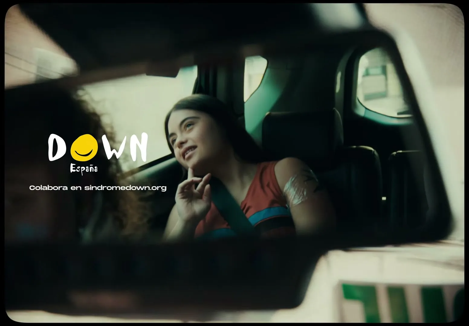 A young woman with long dark hair, wearing a red and blue top, sits in the back seat of a car, looking out the window with a thoughtful expression.
