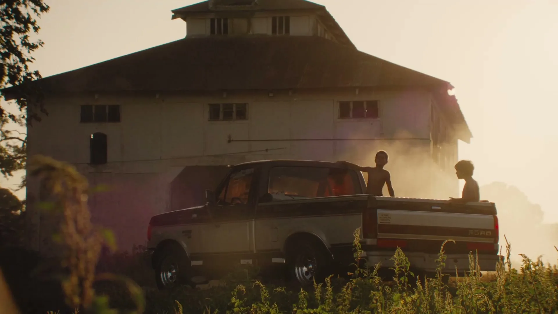 Two children play in the back of a pickup truck, parked in a field in front of a large barn at golden hour.