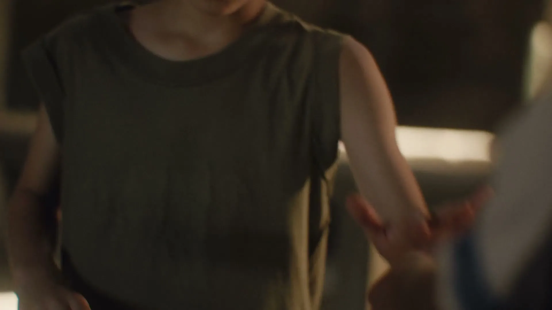 A young person in a green sleeveless shirt gestures with their hands, interacting with someone off-screen in a dimly lit interior.