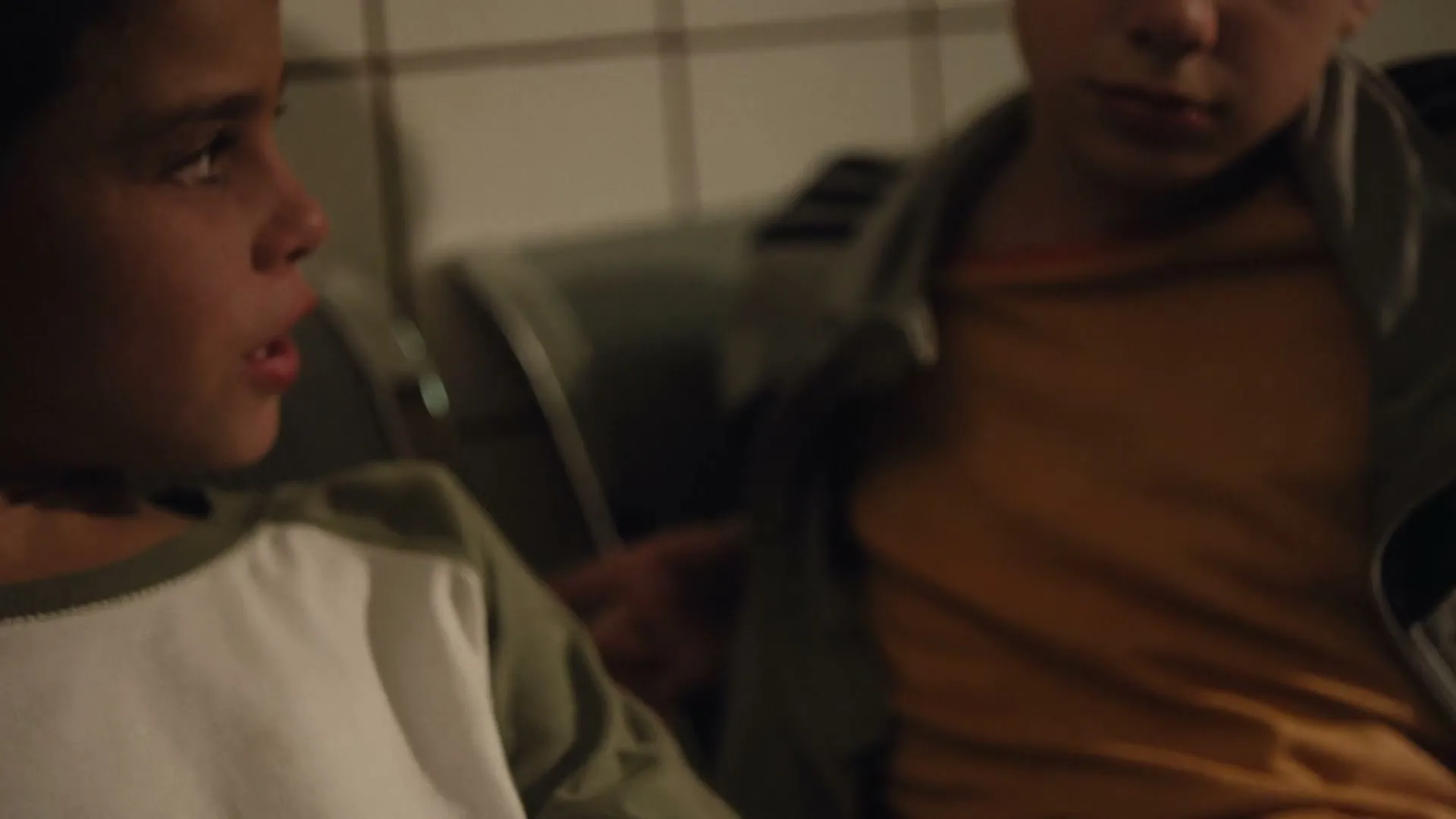 A child applies an ice pack to a scraped knee, then two children sit close together, talking and gesturing in a dimly lit room.