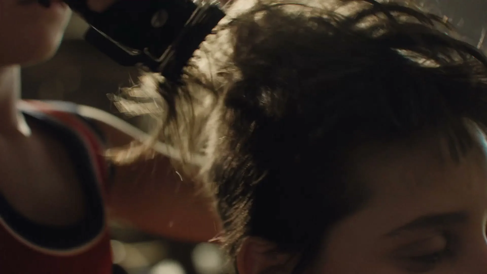 A person in a red tank top shaves another person's head with electric clippers, focusing on the side of their head in a close-up shot.