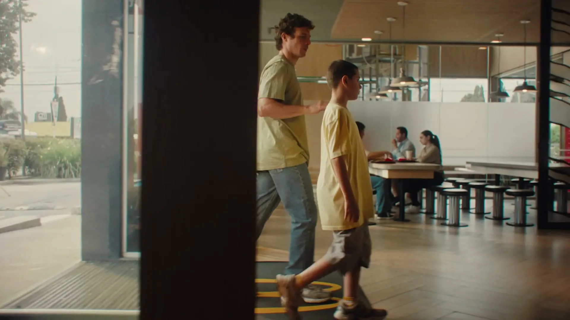 A father and son walk into a McDonald's restaurant, passing other customers seated at tables, with the son looking around curiously.