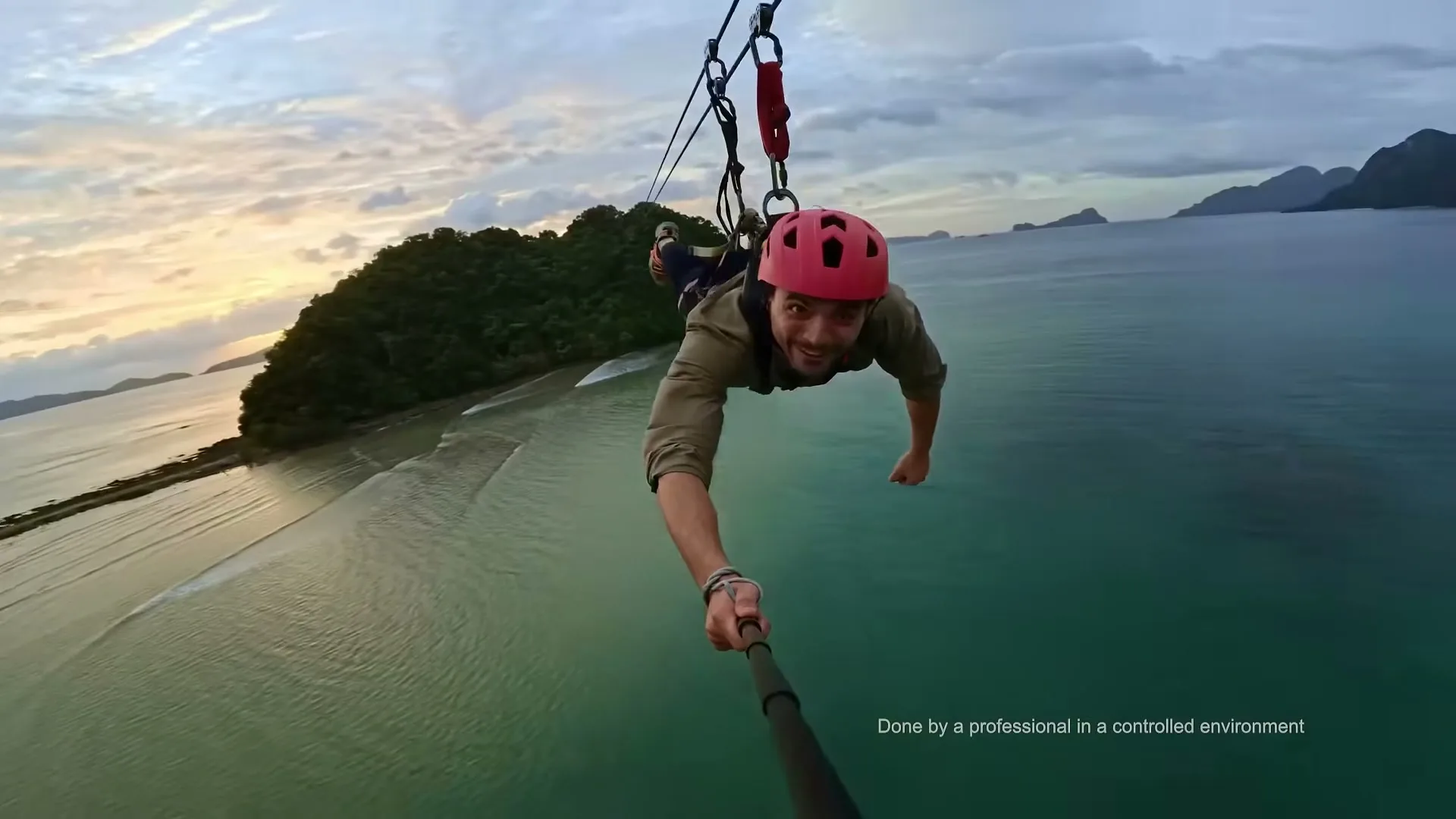 A man in a red helmet and khaki shirt smiles at the camera while ziplining over a tropical beach and ocean at sunset.