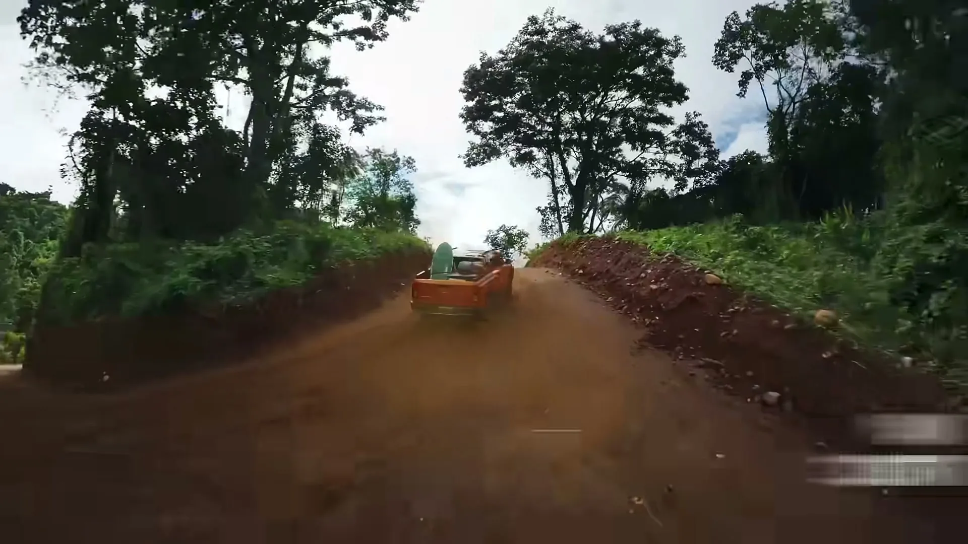 An orange Mitsubishi Triton pickup truck with a surfboard in its bed drives on a dirt road through a lush green jungle under a bright sky.