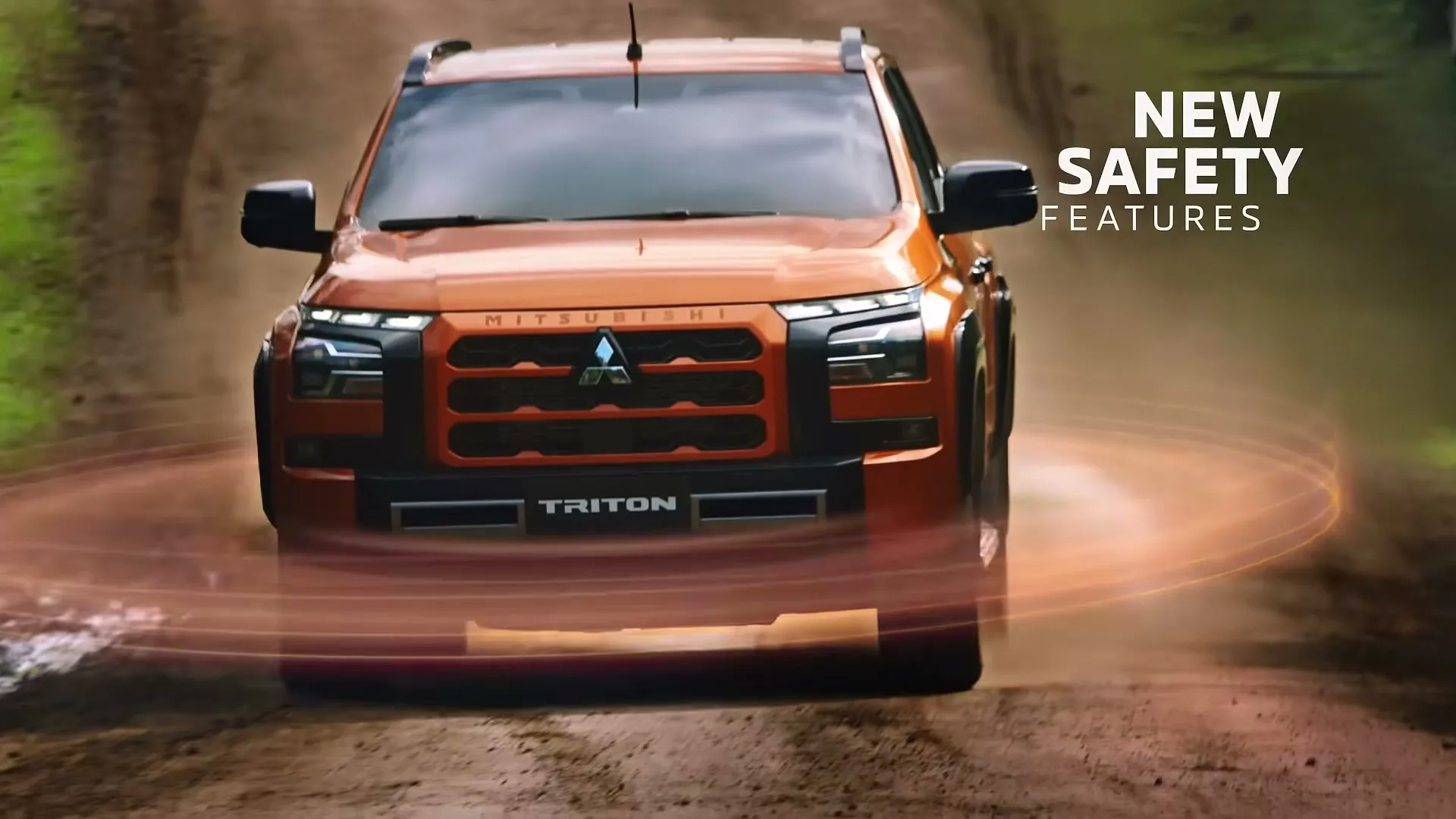 An orange Mitsubishi Triton pickup truck drives towards the camera on a dirt road, with "NEW SAFETY FEATURES" text on the right.