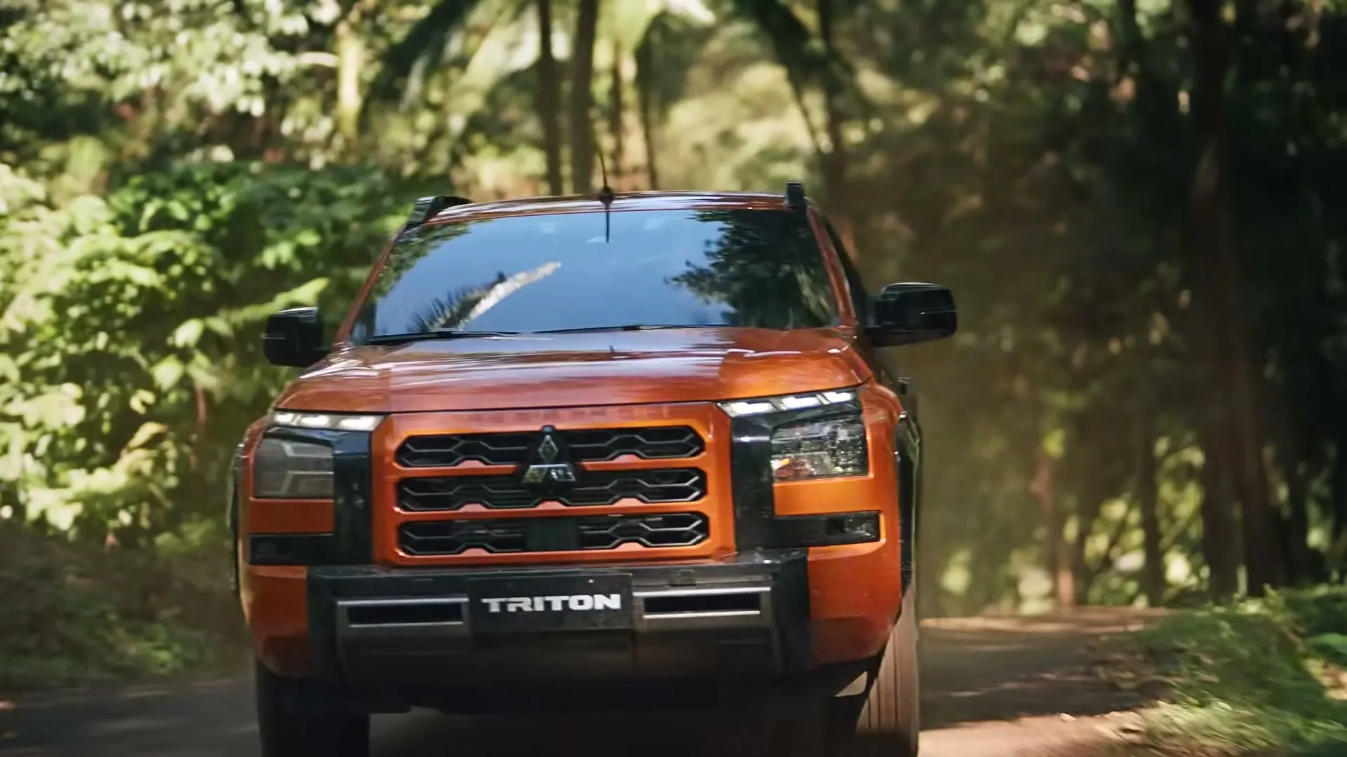 An orange Mitsubishi Triton pickup truck drives towards the camera on a dirt road through a lush green jungle.