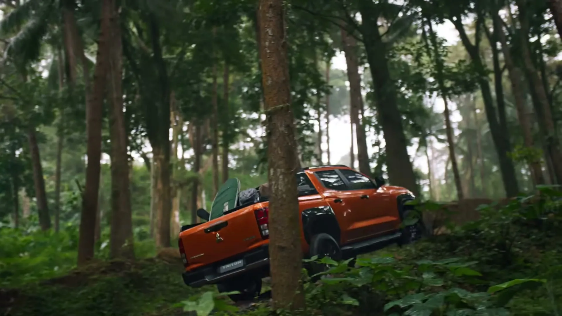 An orange Mitsubishi Triton pickup truck drives uphill through a dense, green forest, showcasing its off-road capability.