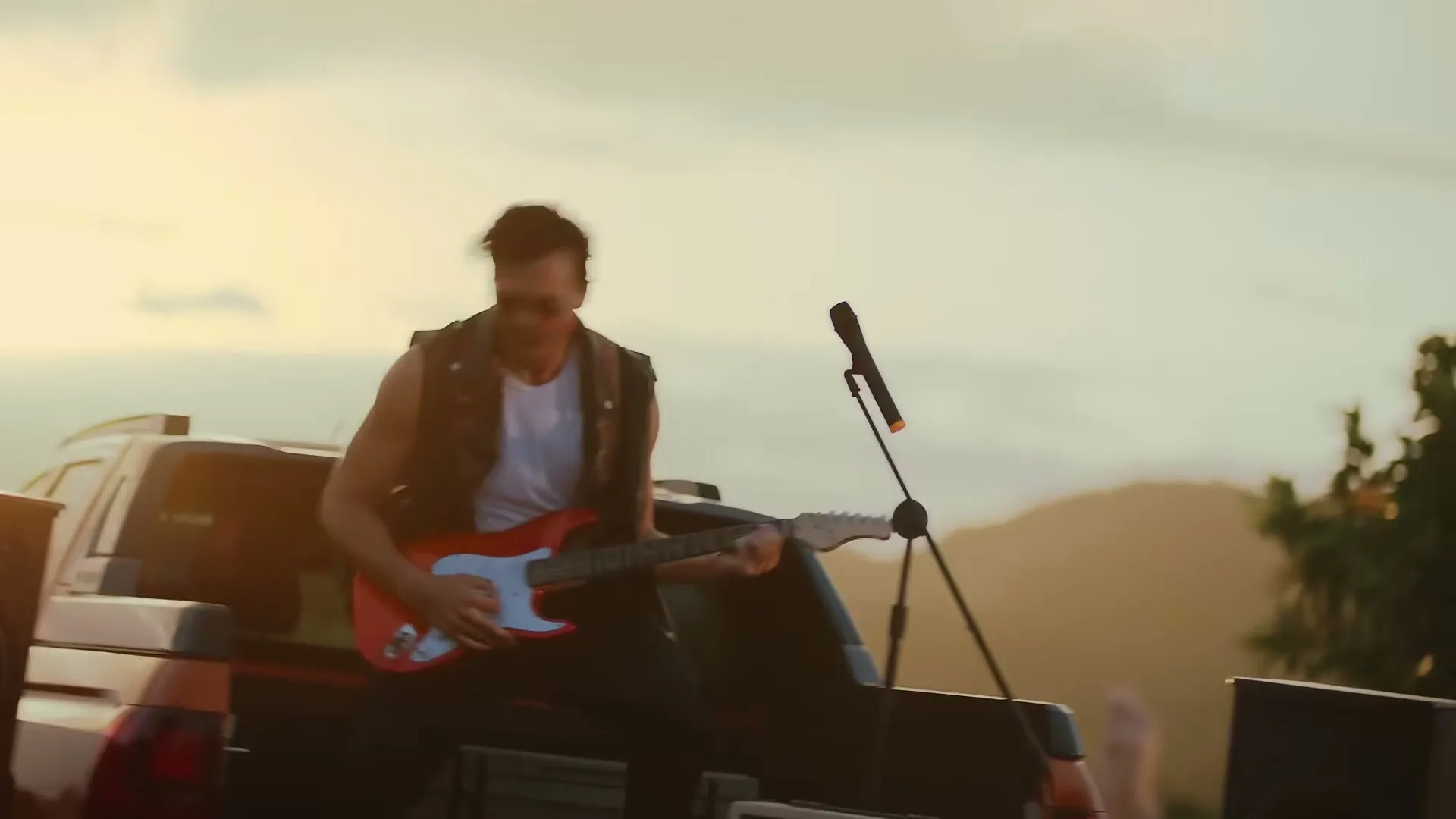 A male musician with sunglasses and a sleeveless leather jacket plays an electric guitar while sitting on the back of a pickup truck at sunset.