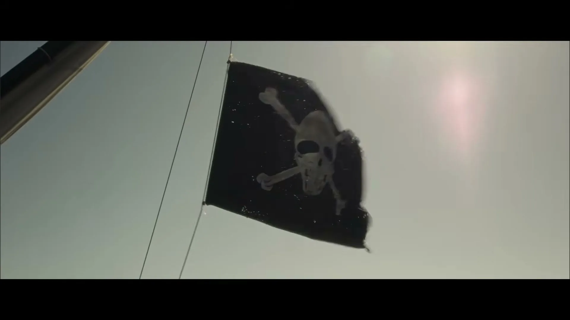 A black pirate flag with a white skull and crossbones waves in the wind against a bright, slightly hazy sky, attached to a ship's mast.