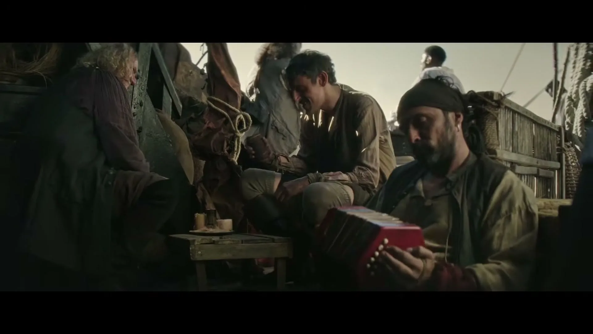 A group of scruffy, old-world sailors are gathered on a ship deck, with one playing an accordion and another laughing.
