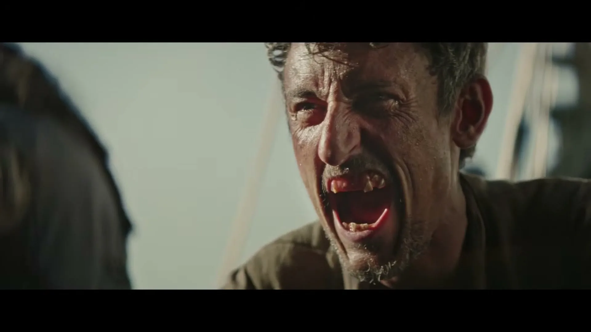 A disheveled man with a beard and missing teeth screams in agony, his face covered in sweat and dirt, on a boat under a bright sky.