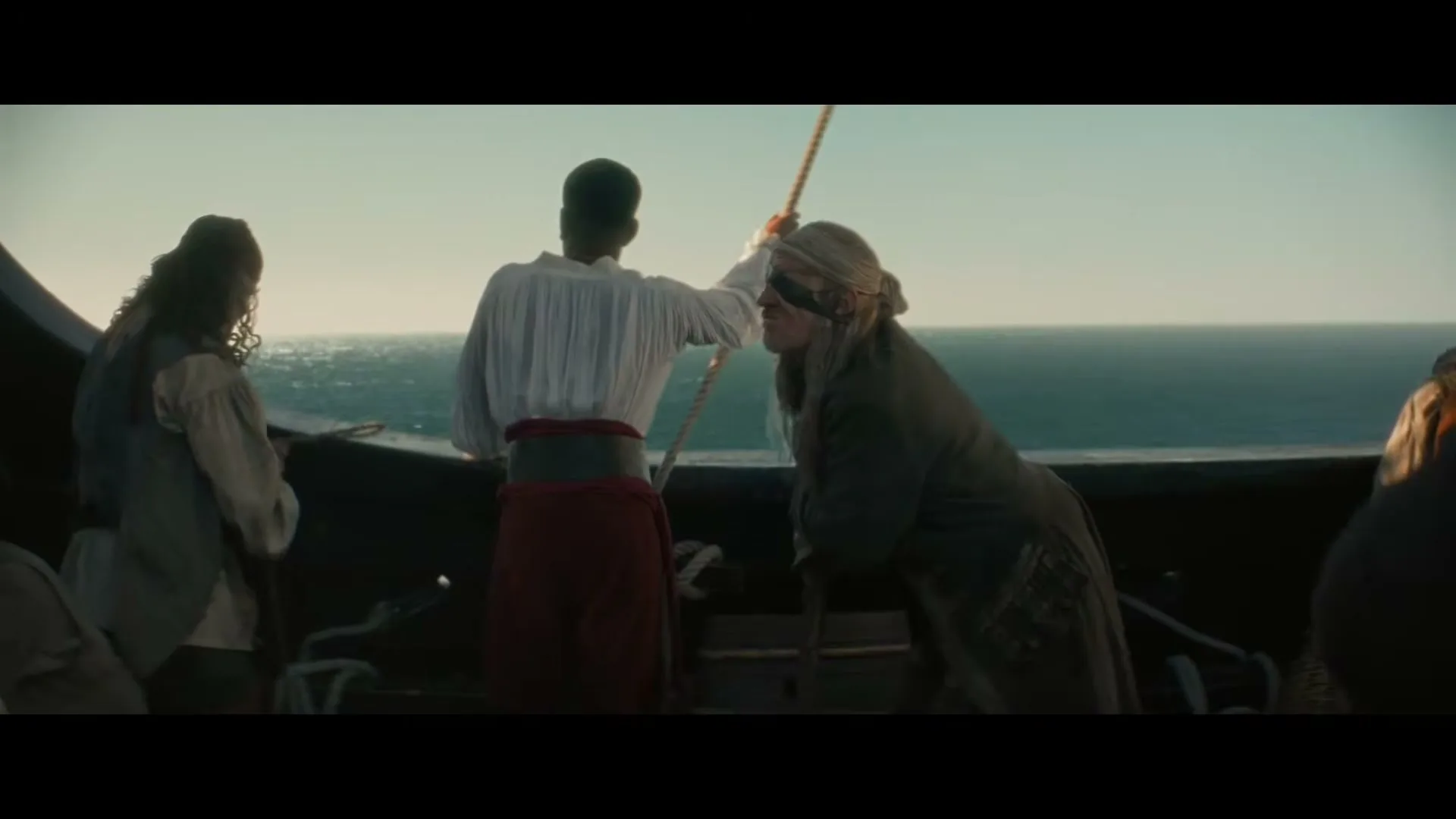 A group of pirates, including one with an eye patch, stand on the deck of a ship, looking out at the ocean under a clear sky.