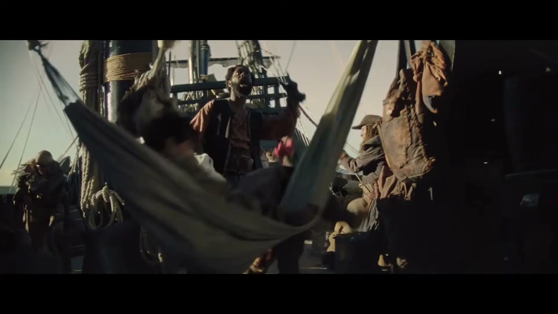 A man in a hammock is violently swung around on the deck of a pirate ship, while other pirates look on in the bright daylight.