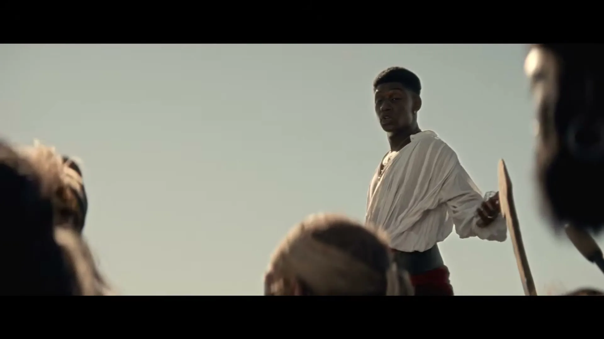 A young Black man in a white tunic and red sash stands among a crowd, holding a wooden shield, under a bright, clear sky.