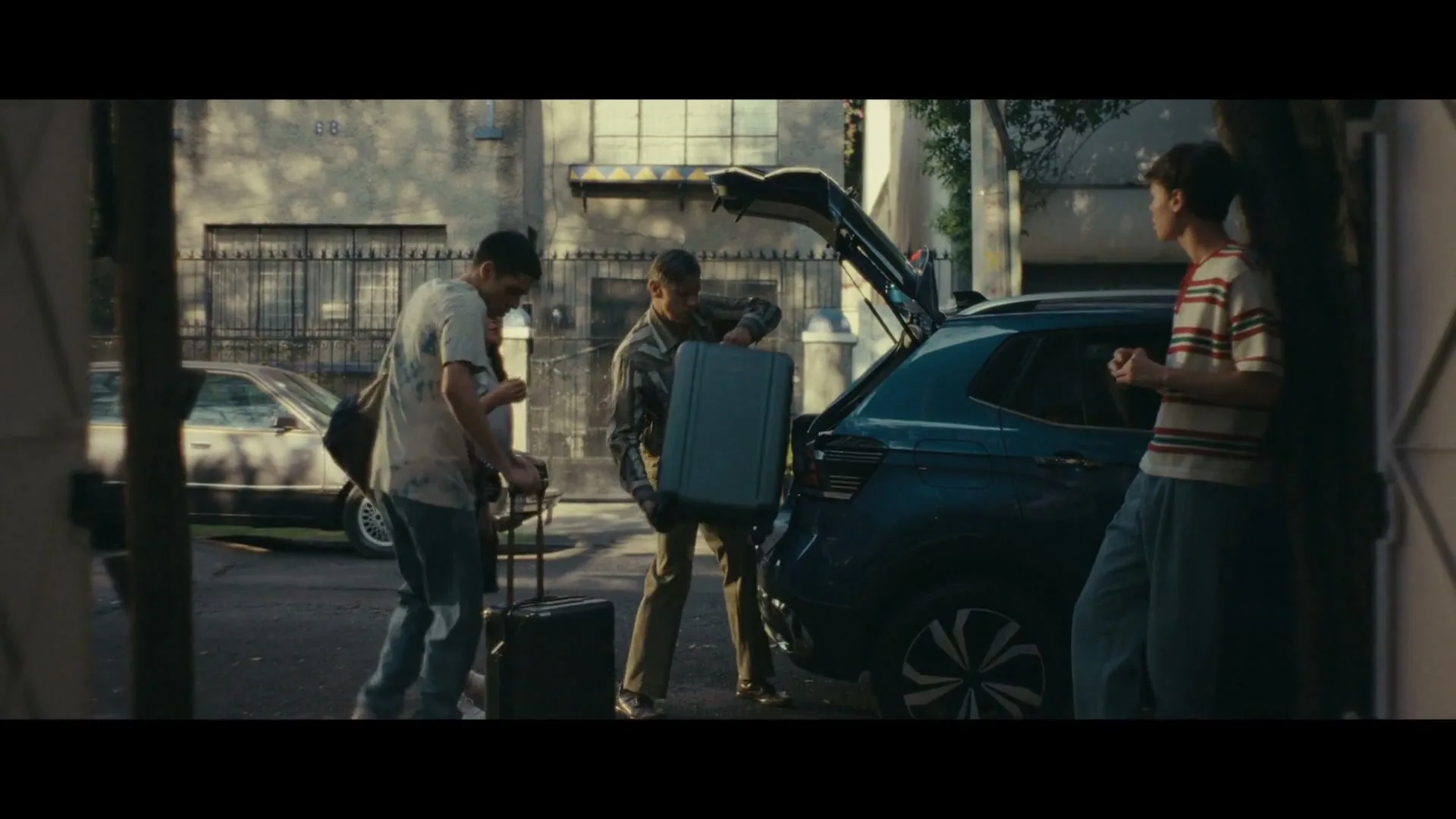 Four young adults, two males and two females, are loading luggage into the trunk of a blue SUV parked on a residential street during the day.