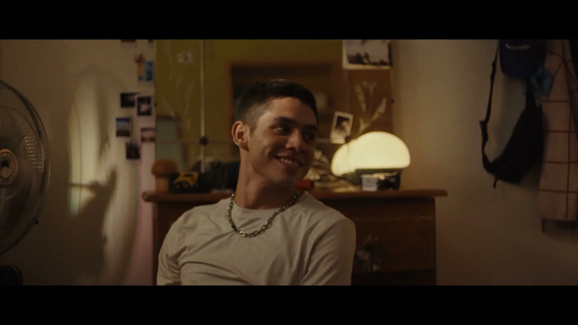 A young man with a short haircut and a silver chain smiles while sitting in a dimly lit bedroom.
