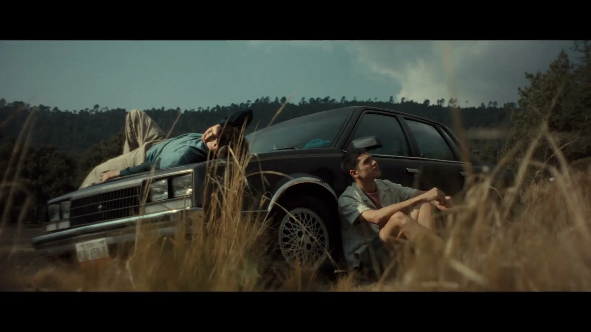 Two young men relax by a broken-down car in a field, one lying on the hood and the other sitting on the ground, looking up at the sky.