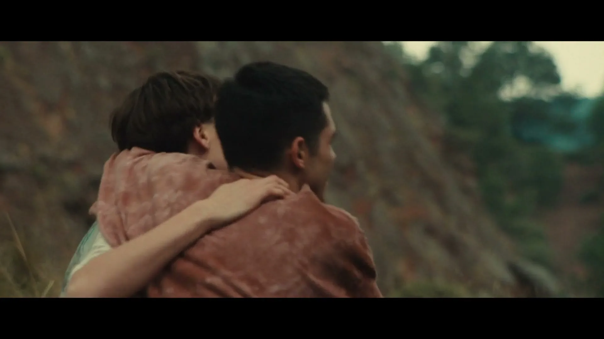 Two young men, one with dark hair and a brown shirt, the other with lighter hair and a white graphic tee, embrace outdoors on a hillside.