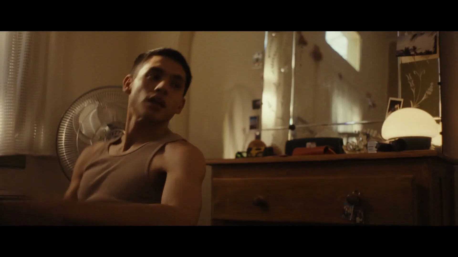 A young man in a tank top sits in a dimly lit room, looking to the side with a thoughtful expression, next to a fan and a dresser.