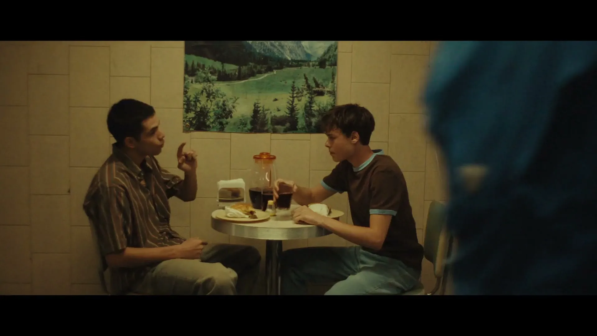 Two young men sit at a small table in a diner, one gesturing while the other drinks from a glass.