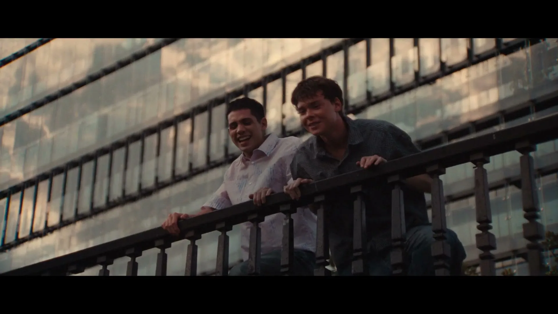 Two young men, one in a white shirt and one in a blue shirt, lean over a railing, smiling and looking up at a modern building with reflective windows.