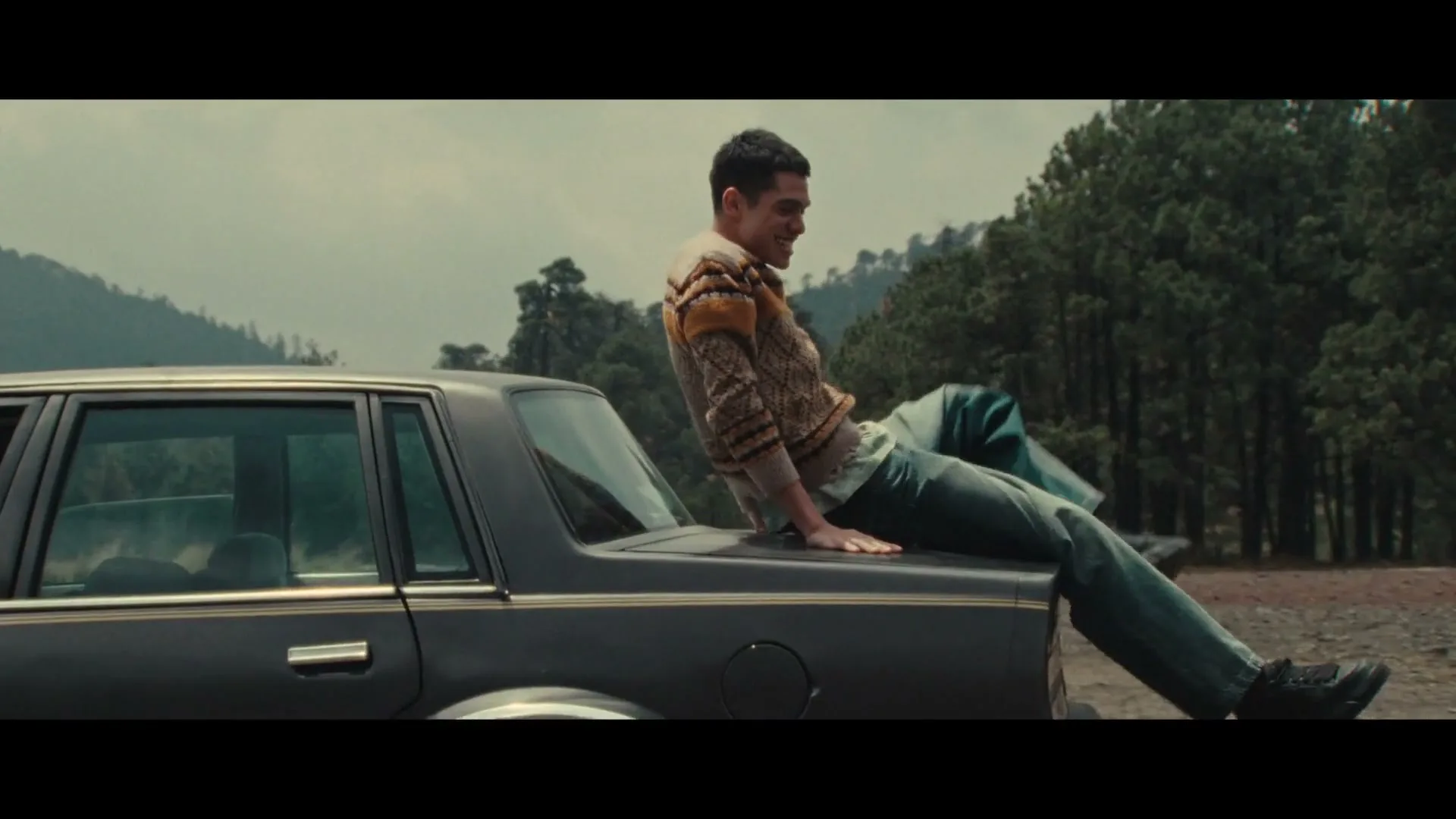 A young man in a striped sweater and jeans climbs onto the trunk of a dark car, smiling, with a forest and mountains in the background.