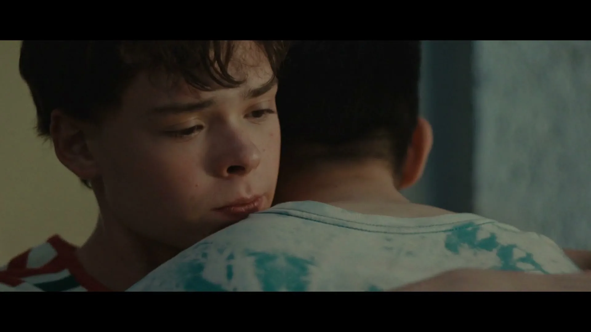 A young boy with short brown hair, wearing a red and white striped shirt, hugs another boy from behind, whispering into his ear in a dimly lit room.