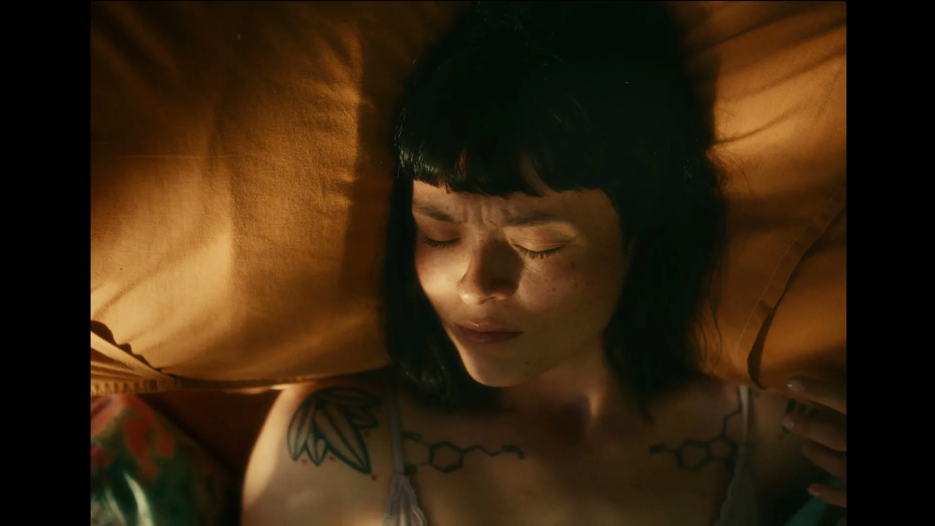 A young woman with dark hair and tattoos lies in bed, eyes closed, as she slowly wakes up in a warm, dimly lit room.