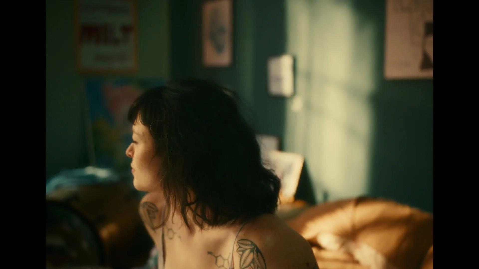 A young woman with dark hair and tattoos wakes up in bed, stretching and looking around her messy room in the morning light.