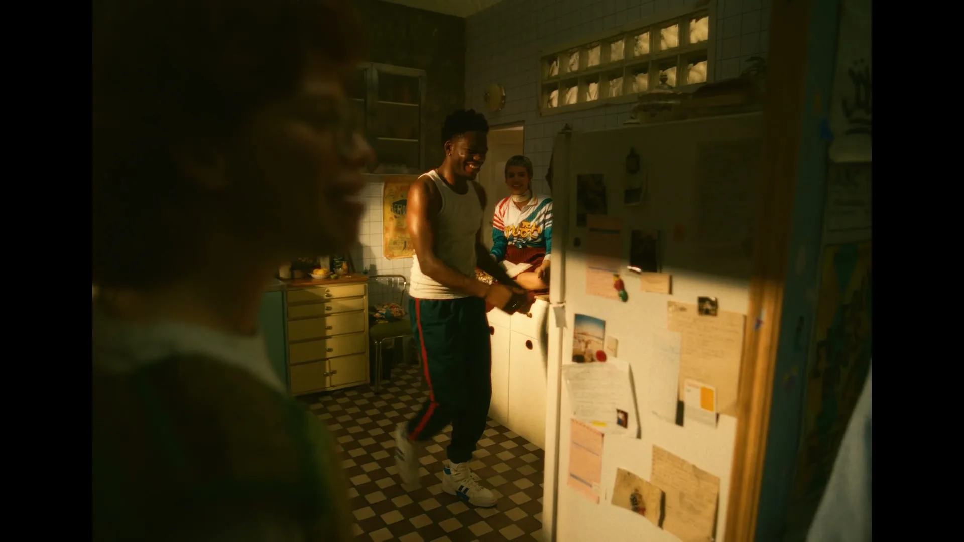 A diverse group of friends gathers in a dimly lit kitchen, with one man dancing playfully while another sits on the counter.