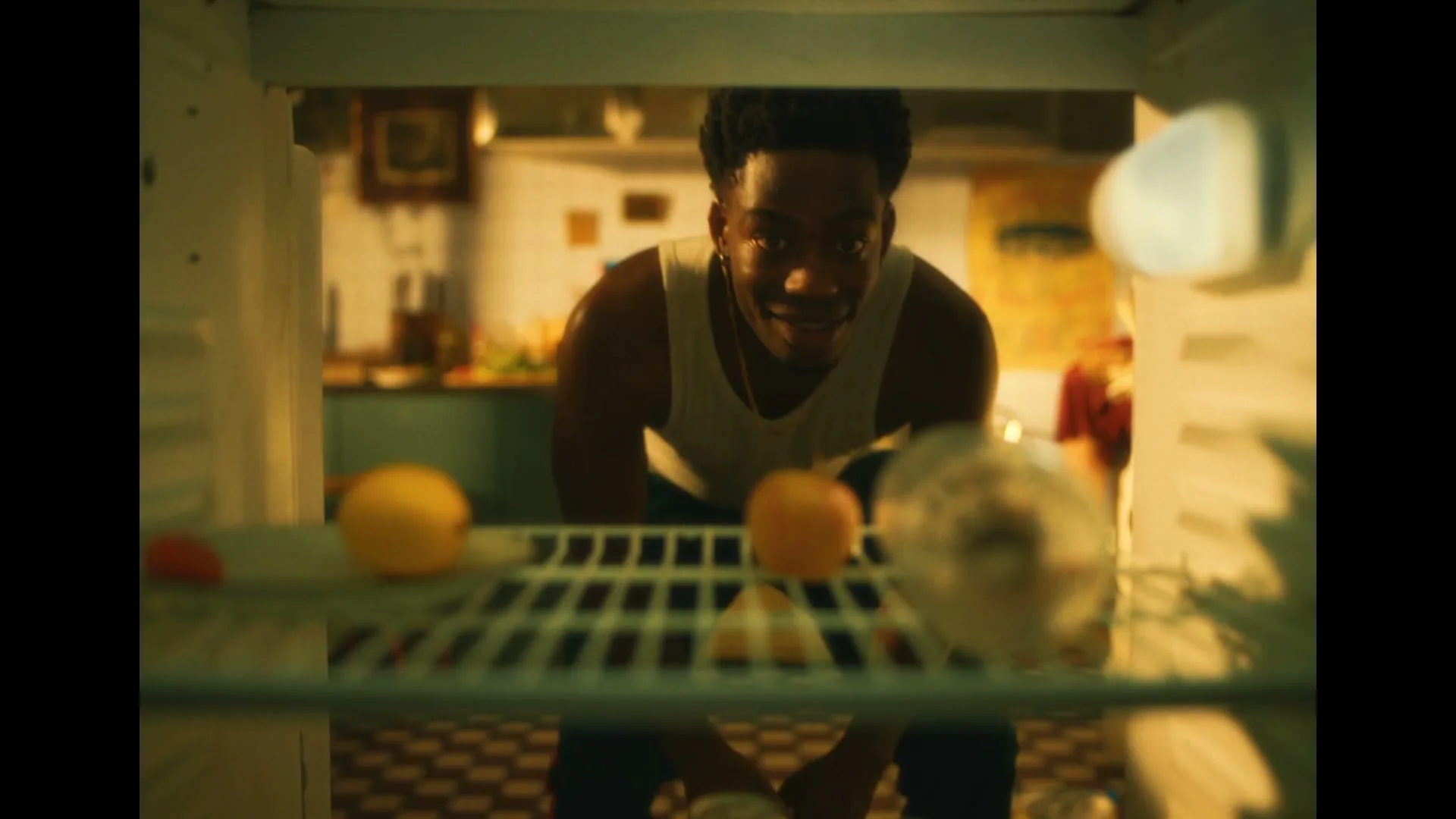 A young Black man in a white tank top looks into an empty fridge, searching for something to drink in a warm-toned kitchen.