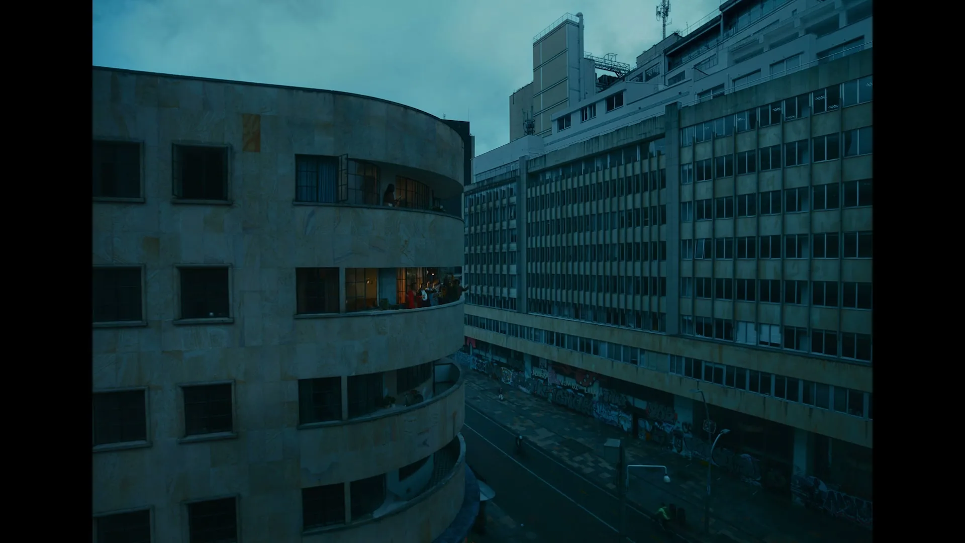 A wide shot of a city street at dusk, with a curved building on the left and a rectangular building on the right. People are visible in the windows of the curved building.