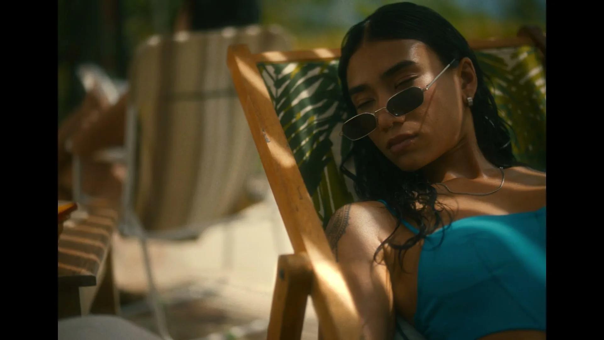A young woman with dark hair and sunglasses relaxes in a patterned deck chair, eyes closed, on a sunny day outdoors.