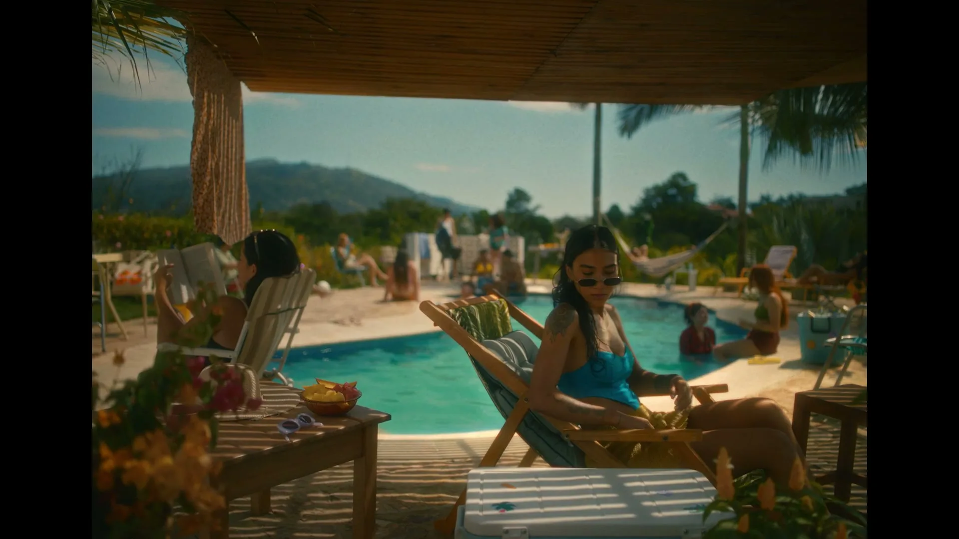 A young woman in a blue swimsuit and sunglasses relaxes in a lounge chair by a pool, opening a cooler at a lively outdoor party.