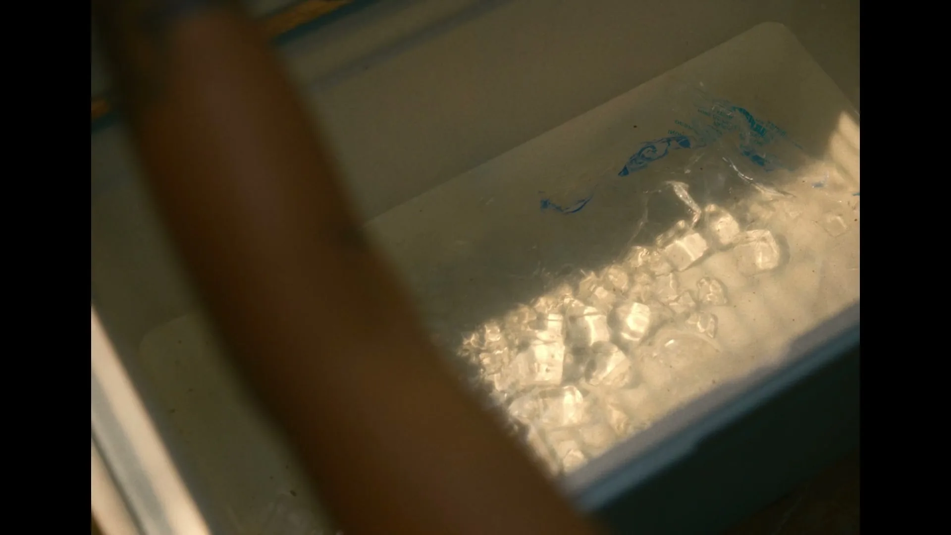 A person's arm reaches into a cooler filled with ice and water, with sunlight reflecting on the surface.