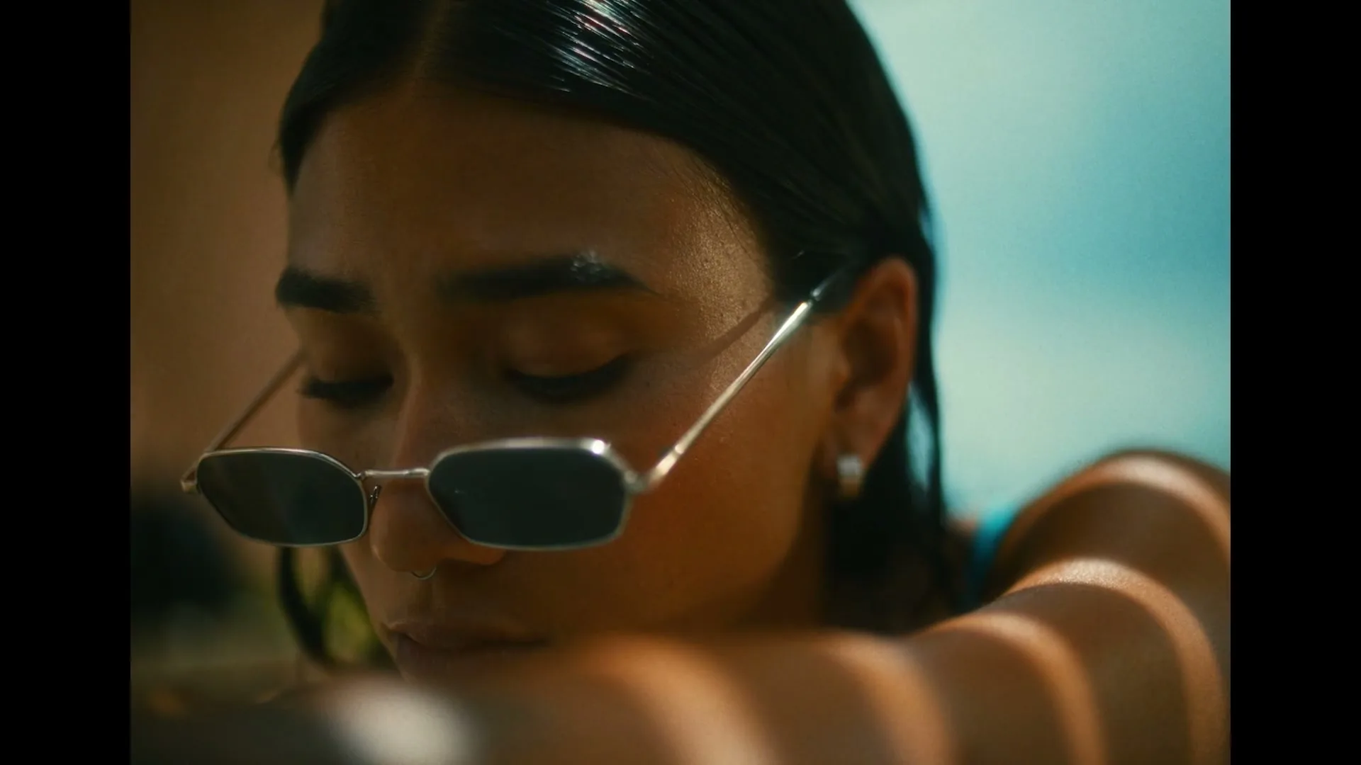 A young woman with wet hair and sunglasses rests her head on her arm, eyes closed, in a sun-drenched outdoor setting.