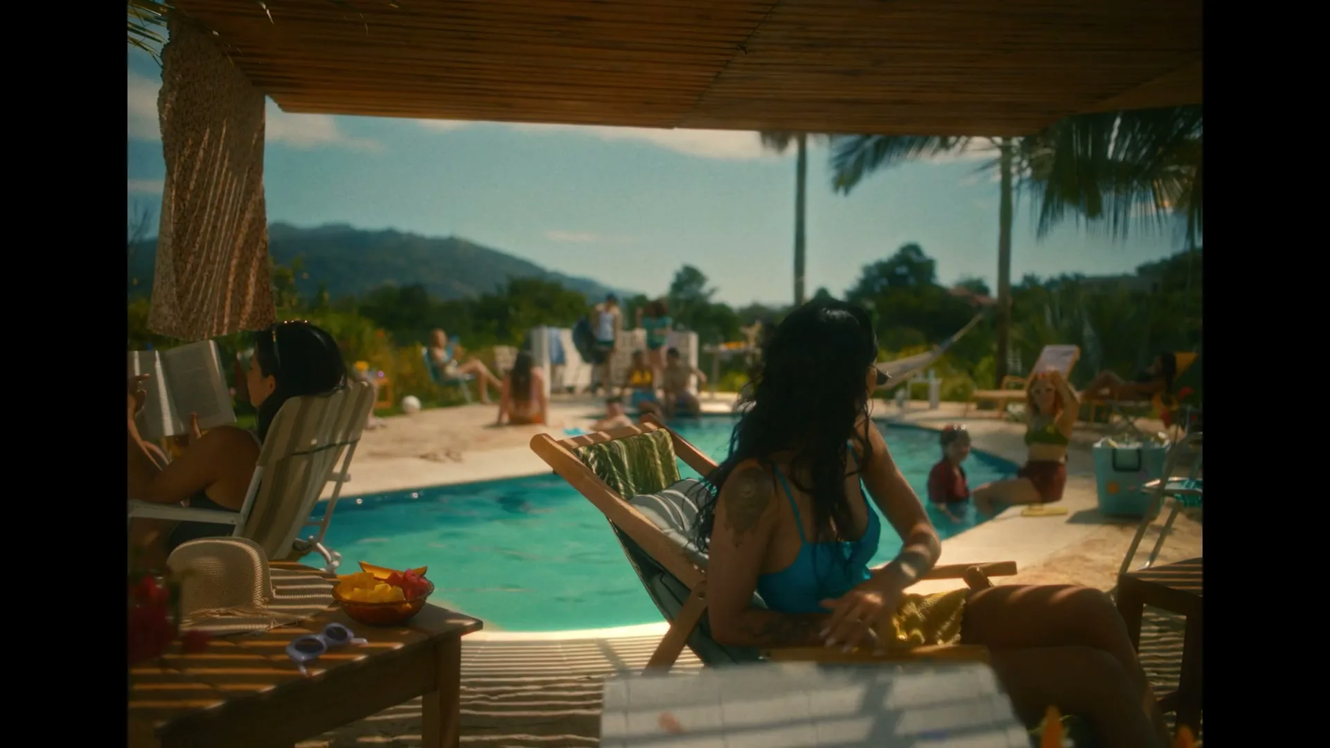 A woman in a blue bikini sits under a shaded patio next to a pool, looking over her shoulder at a lively pool party with many people.