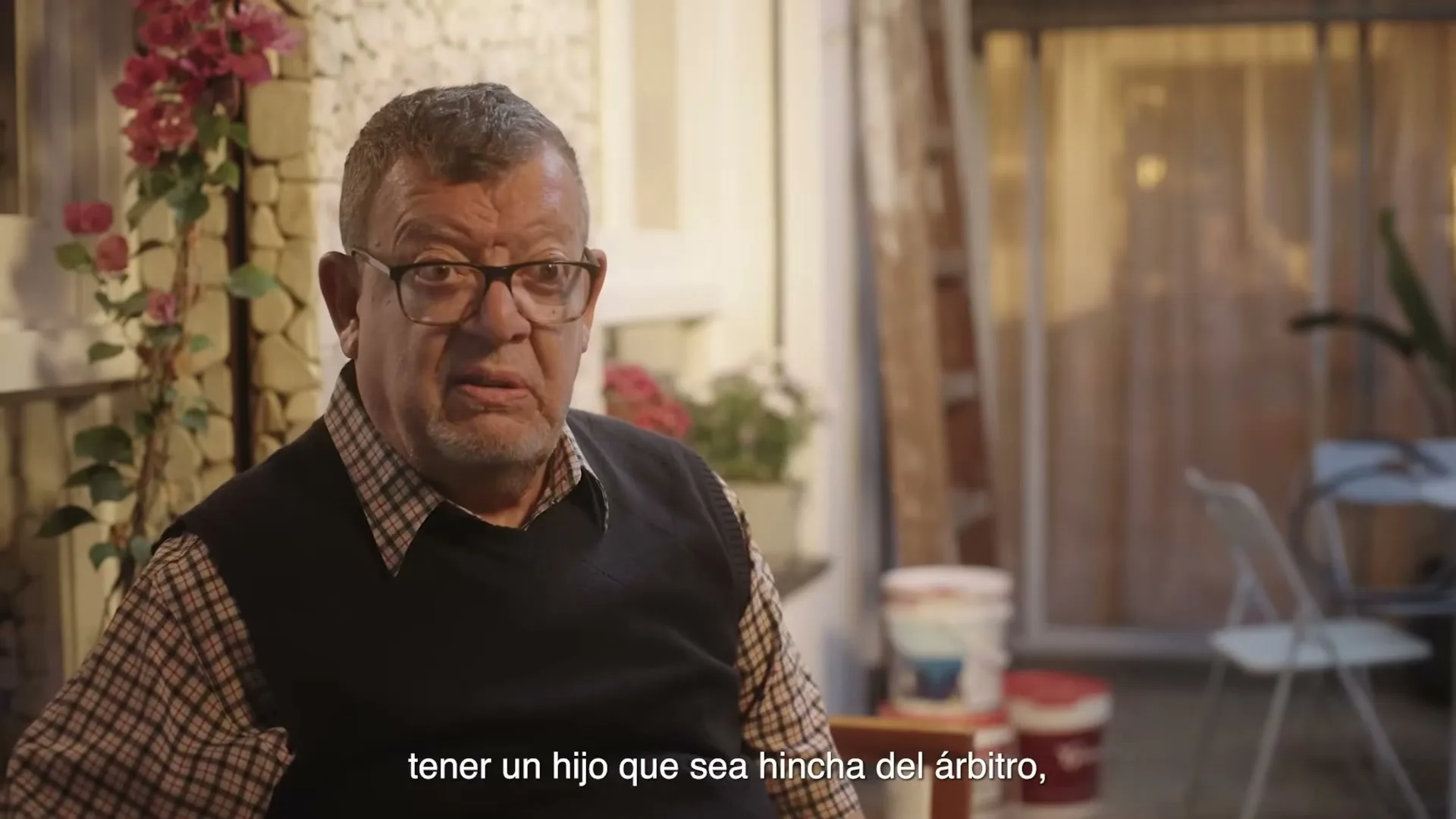 A middle-aged man with glasses and a plaid shirt speaks directly to the camera in a casual outdoor setting, with plants and a patio in the background.