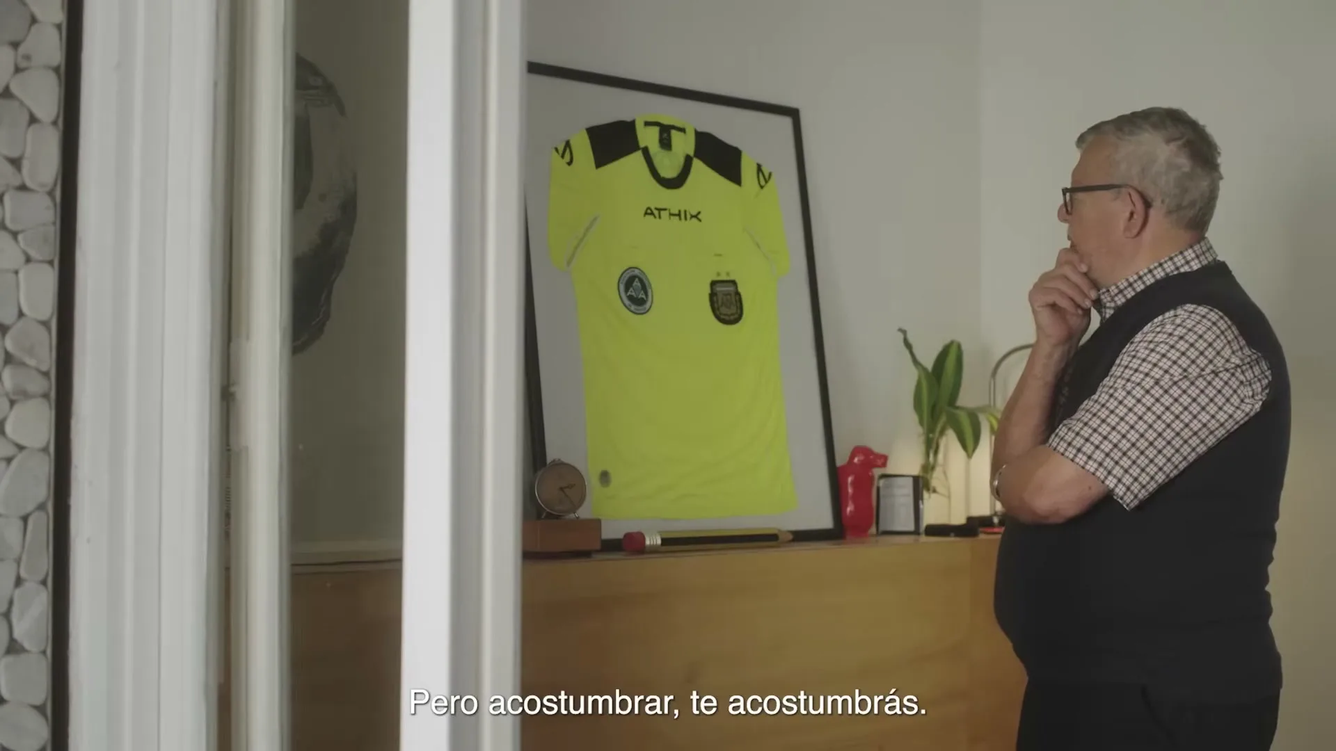 An older man in glasses and a vest looks thoughtfully at a framed, bright yellow soccer jersey on a wooden dresser in a home.