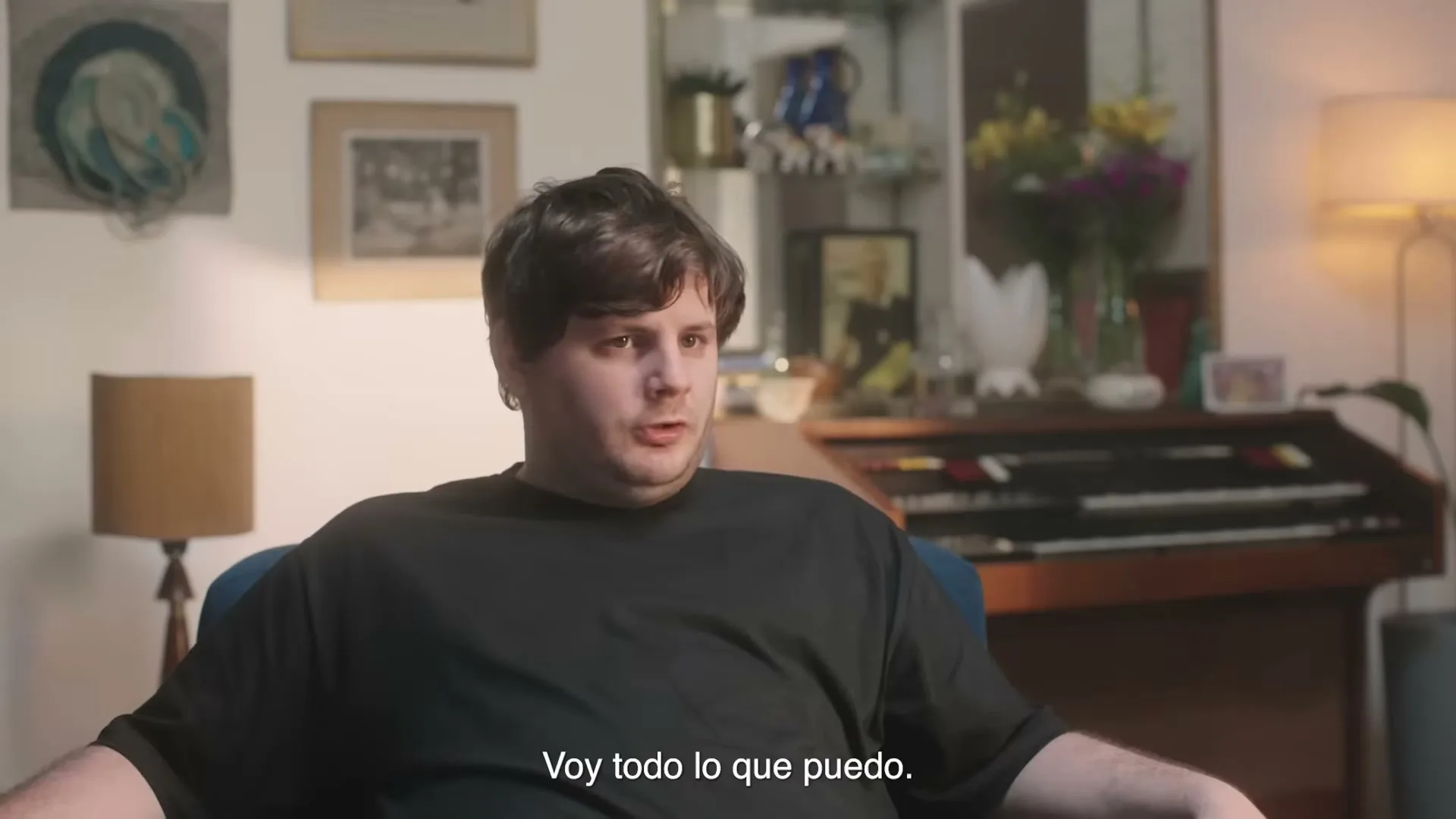 A man in his 20s with dark hair and a black t-shirt sits in a blue armchair, looking forward with a neutral expression in a cozy living room.