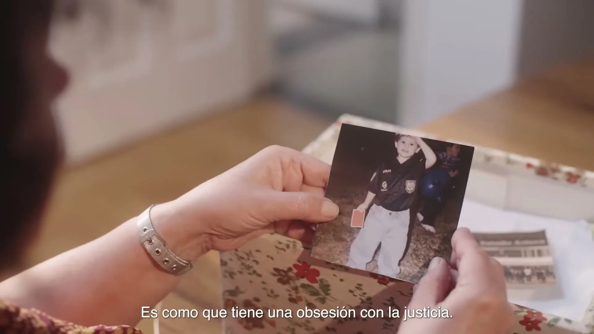 A woman with a silver watch on her wrist looks at an old photo of a child in a soccer uniform, holding a red card, in a floral box.