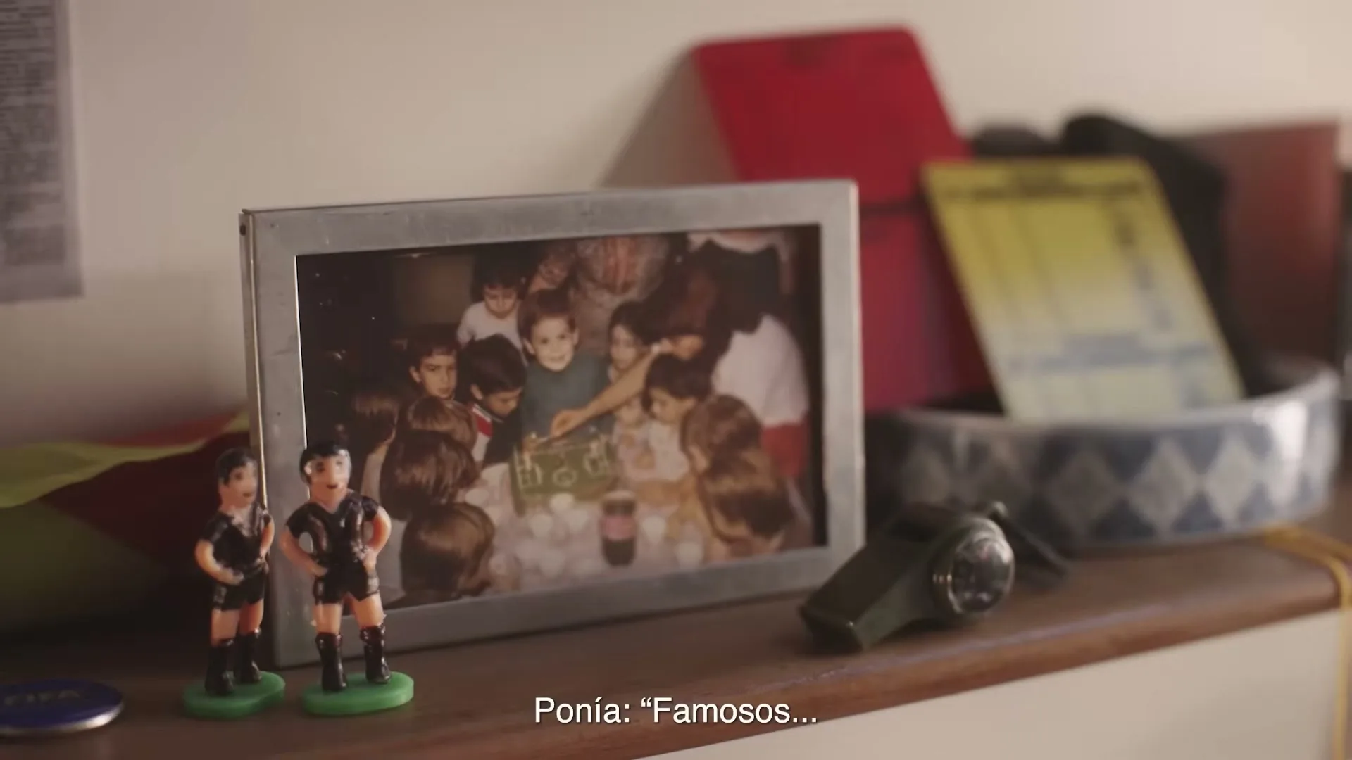 A framed photo of children at a birthday party sits on a wooden shelf next to two small soccer player figurines and a whistle.