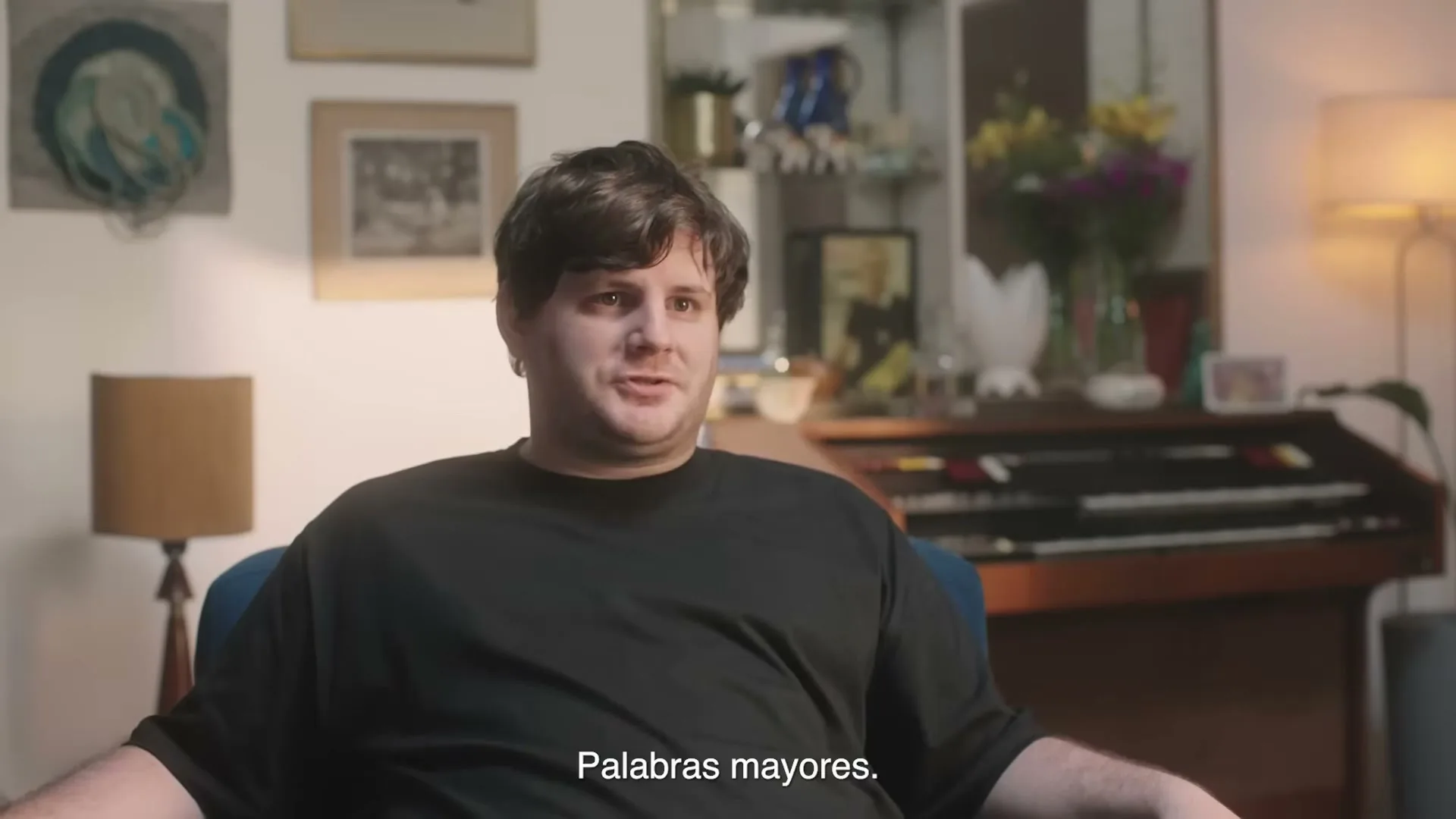 A young man with dark hair and a black t-shirt sits on a blue armchair in a living room, looking slightly to the right and talking.