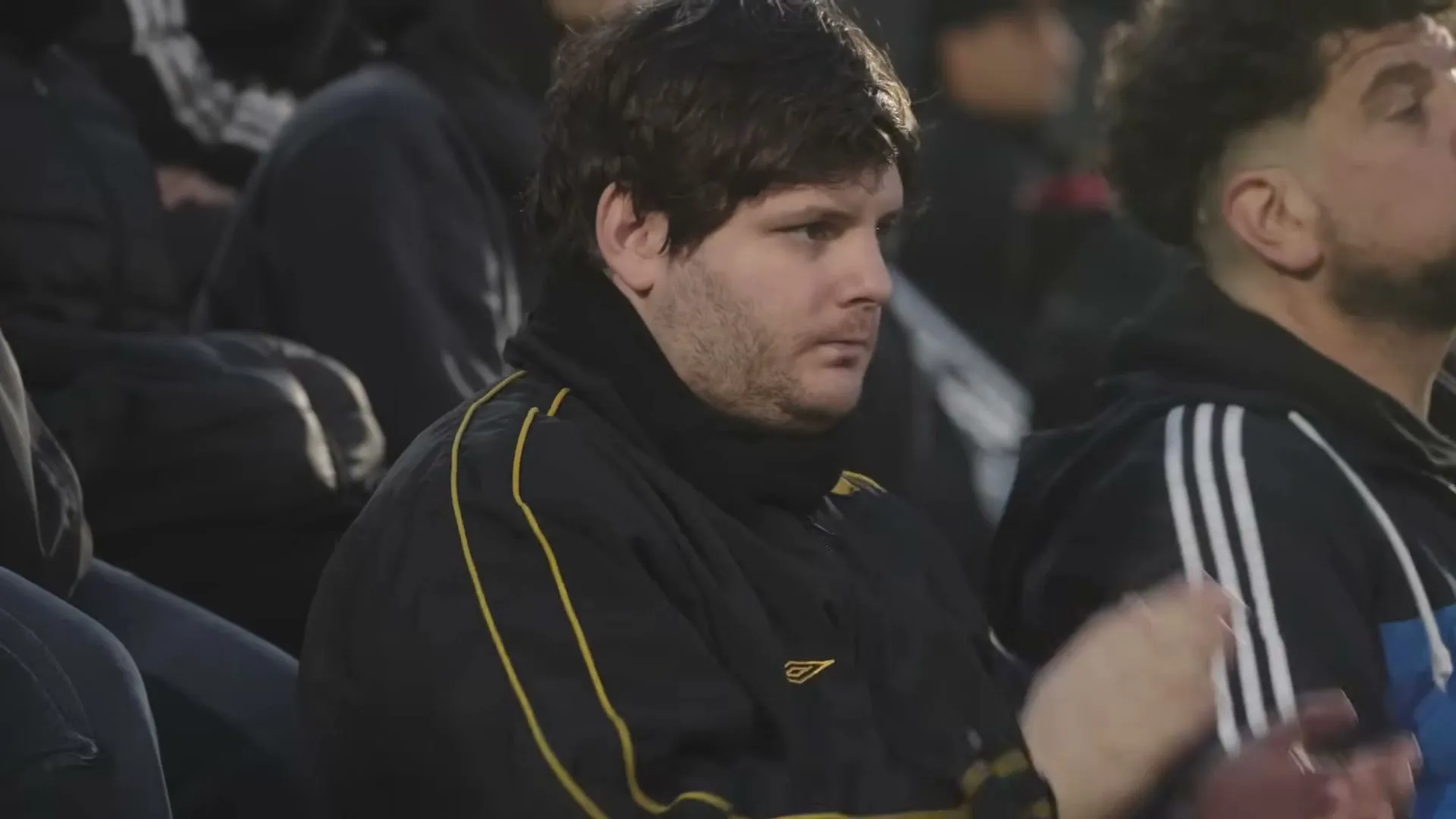A male soccer fan in his 30s, wearing a black and yellow jacket, looks around and then claps in a stadium.
