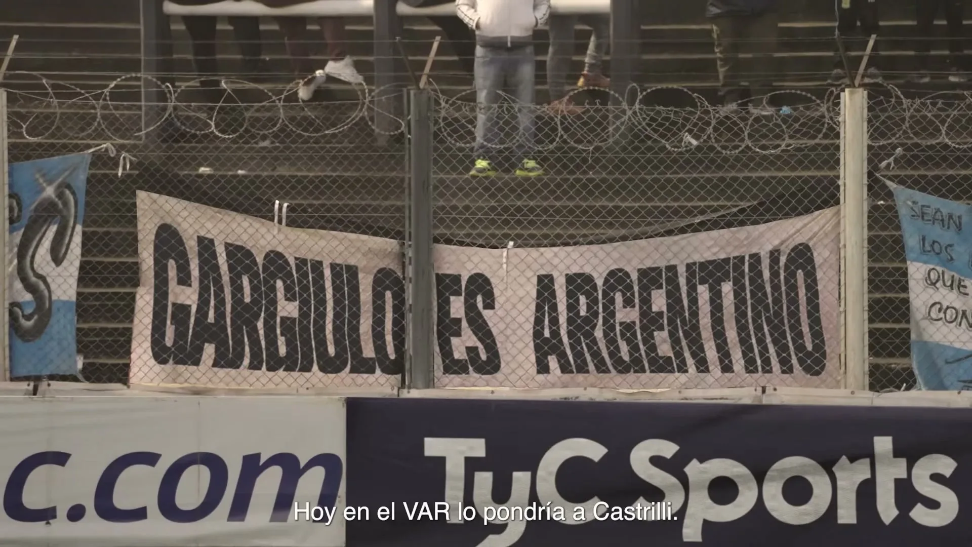 A banner with the text "GARGIULO ES ARGENTINO" is displayed at a stadium, with people standing behind a fence.