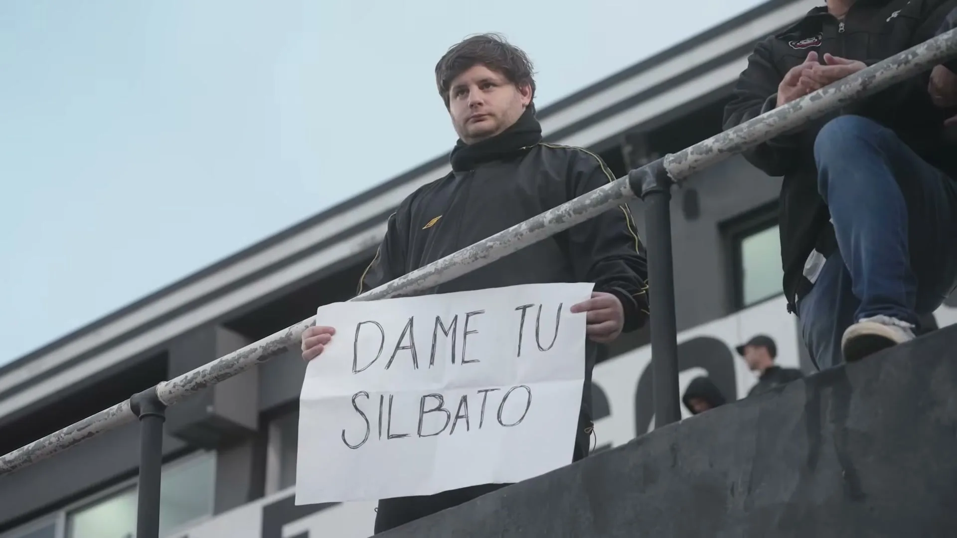 A male soccer fan in a black and yellow tracksuit holds up a sign that says "DAME TU SILBATO" (Give me your whistle) in a stadium.