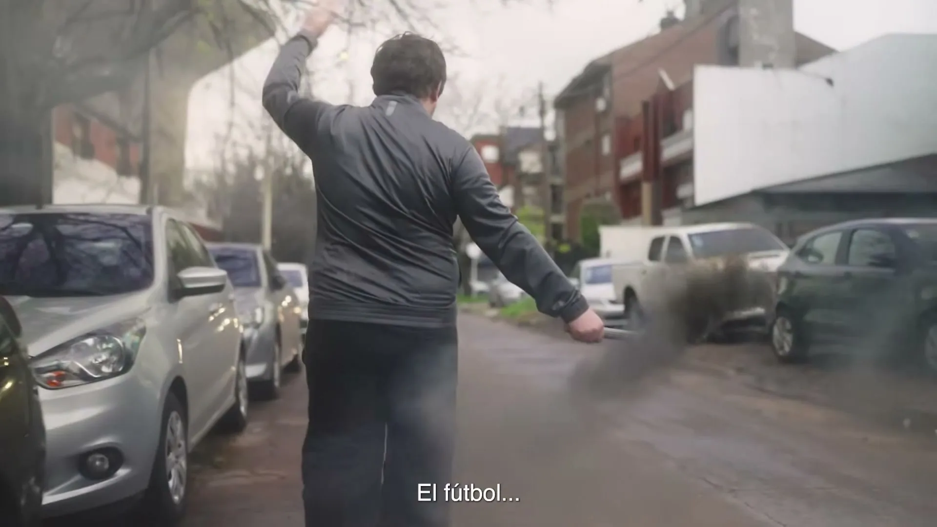 A man in a grey jacket and black pants walks away from the camera on a wet street, raising his arms as if conducting, with parked cars and buildings in the background.