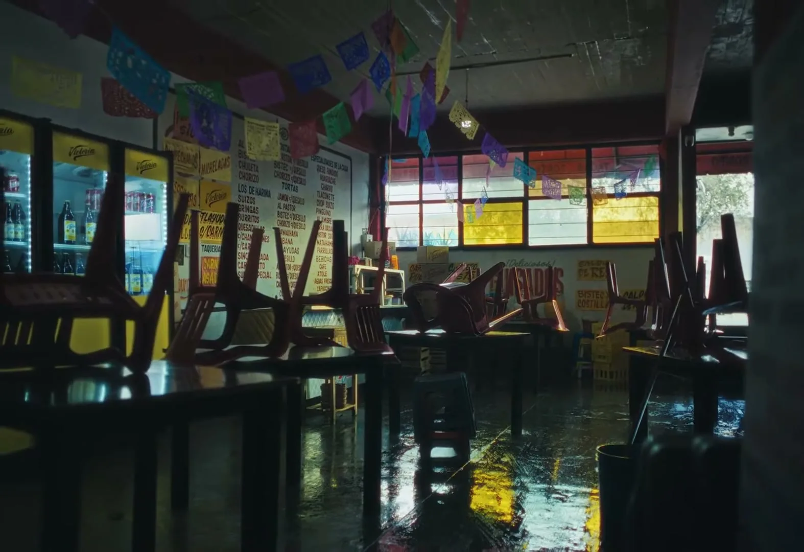 An empty taqueria with chairs stacked on tables, colorful papel picado hanging, and light reflecting on the wet floor from windows.