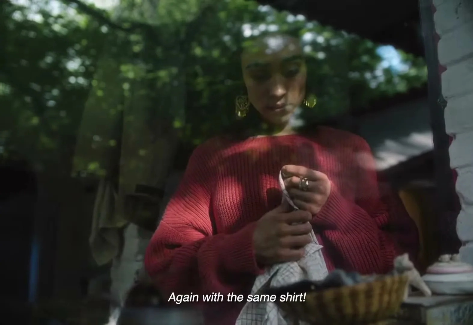 A young woman with dark hair and a red sweater is seen through a window, mending a piece of cloth with a needle and thread.