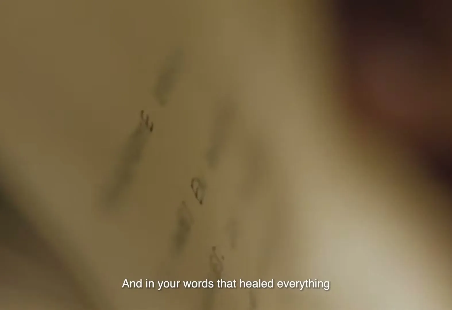 A close-up shot shows handwritten text on an old, yellowed page, with a hand visible in the blurry background, as the camera slowly moves across the page.